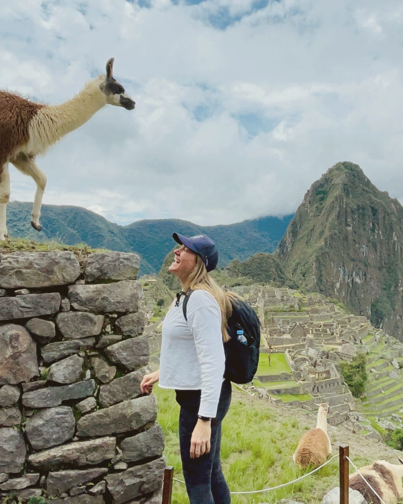 Patricia smiling up at a llama standing on a stone wall at Machu Picchu